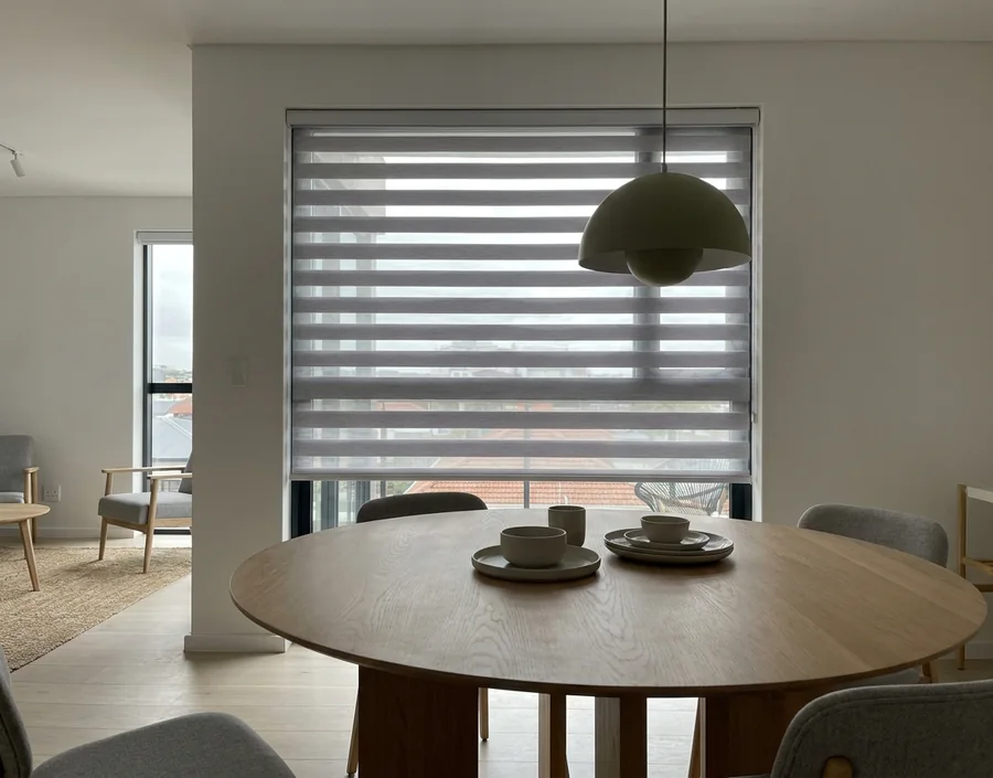 Grey-and-white day/night blind lowered behind a round oak dining table in a Sandown apartment, striped light on the tabletop